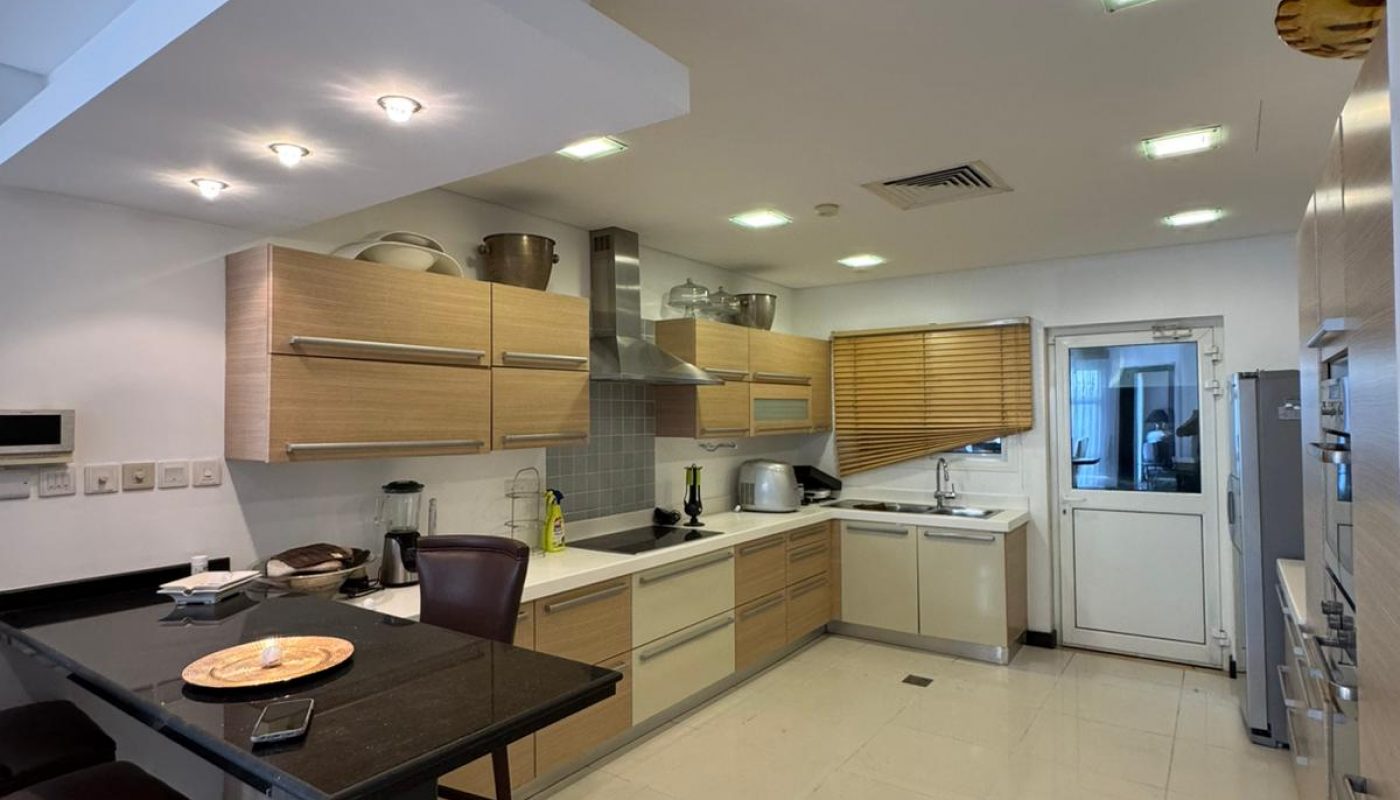 Modern kitchen with wooden cabinets, black countertop, and stainless steel appliances. Ceiling lights illuminate the room. A chair and a table are in the foreground.