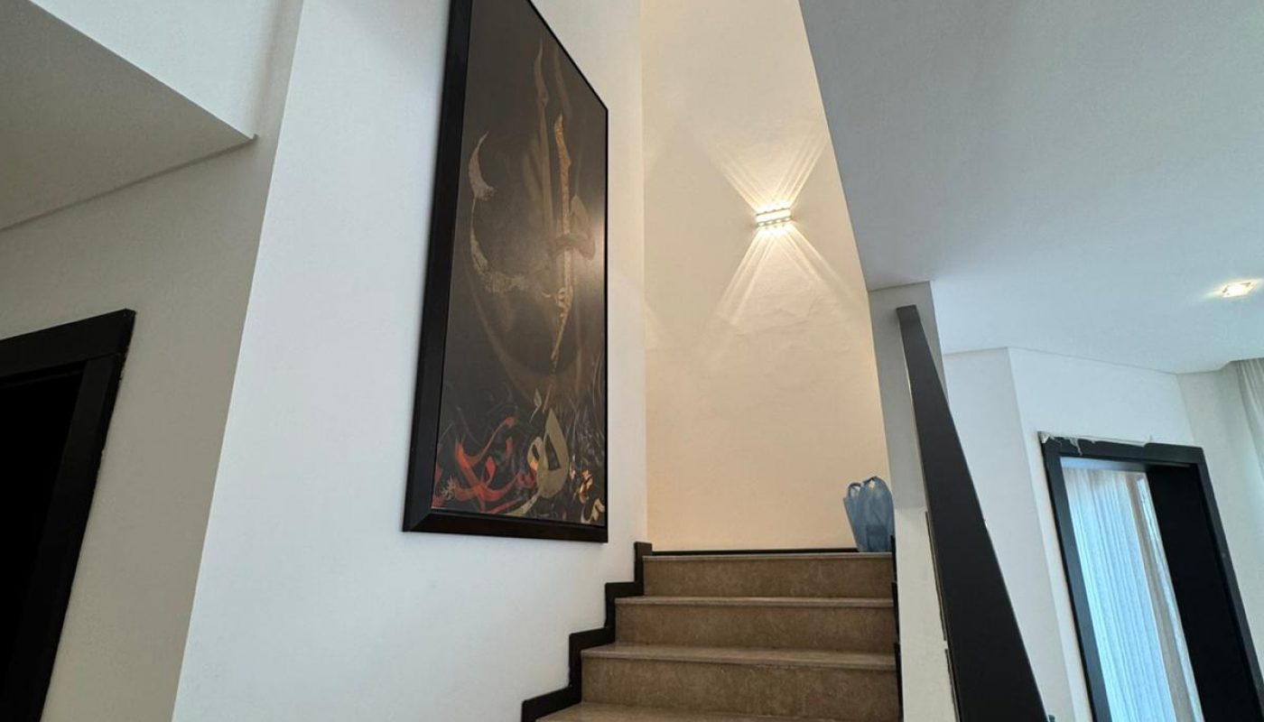 View of a stairway with beige steps, a large framed artwork, and a wall light above. A blue decorative vase is placed near the landing.