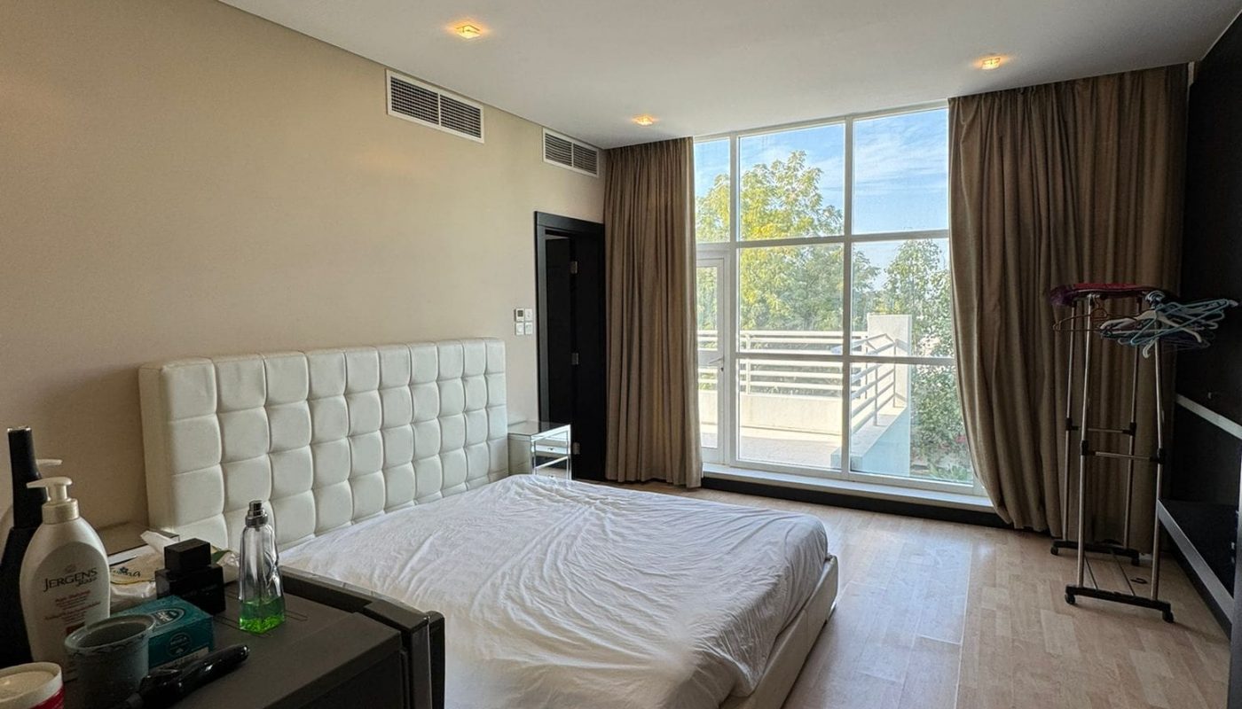 A bedroom with a large window, beige walls, and wood flooring. It has a bed with a white headboard, a dresser, and curtains partially open to a balcony view.