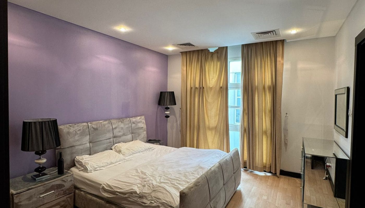 A bedroom with a double bed, lavender accent wall, two bedside tables with lamps, large windows with curtains, a TV, and a desk.