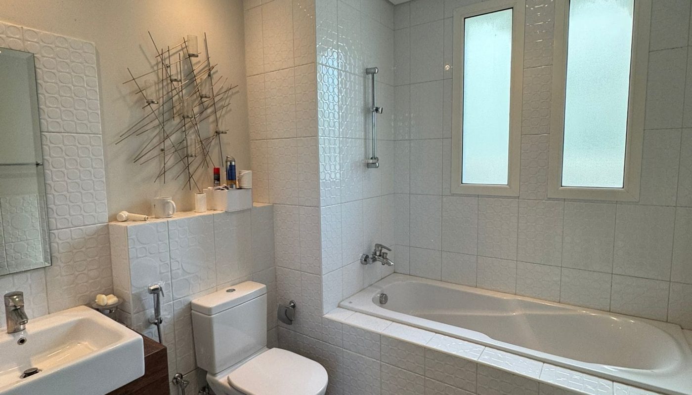 A modern bathroom with a bathtub, toilet, sink, and decorative wall art. The walls and floors are tiled, and there are frosted windows above the bathtub.
