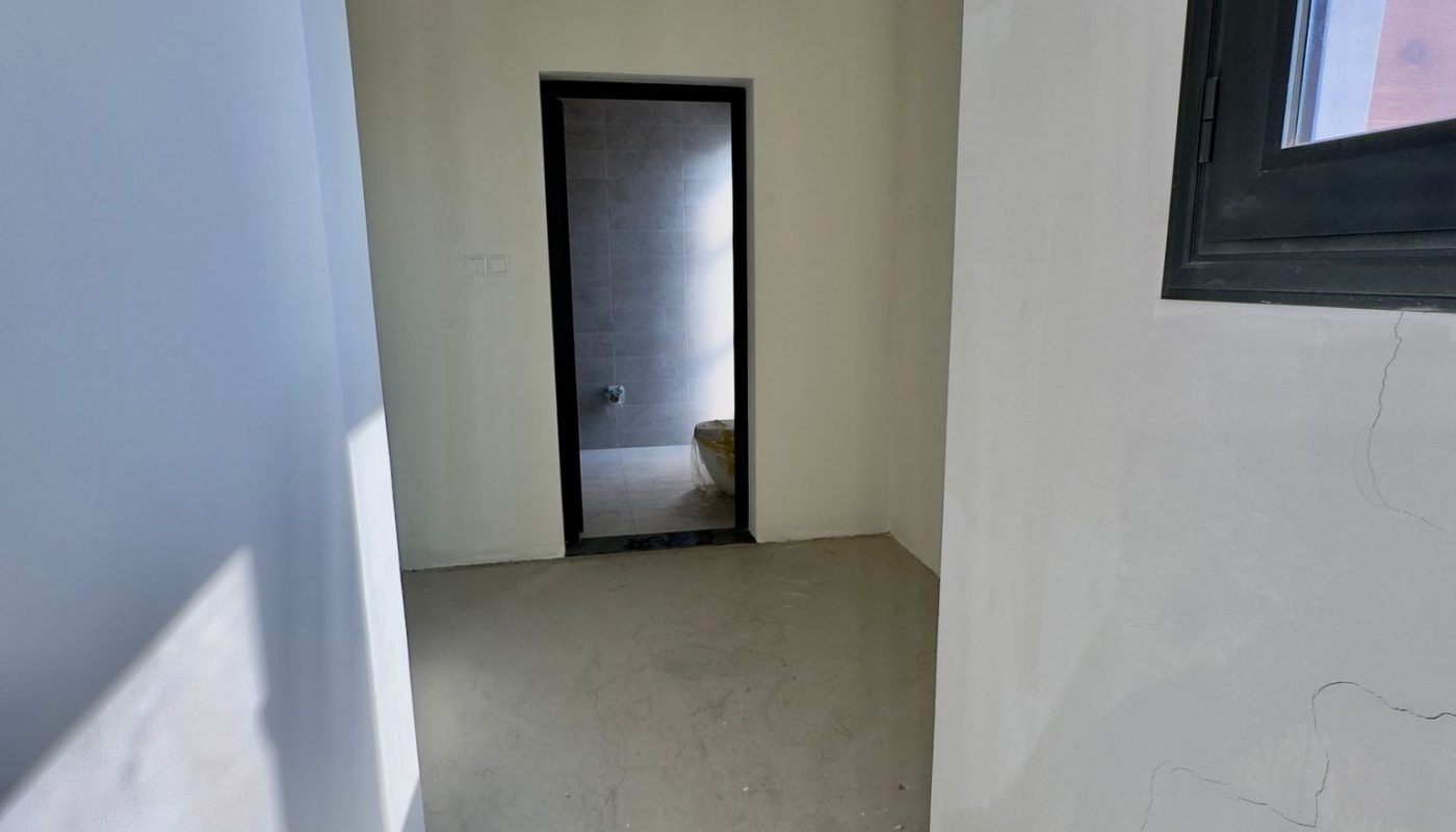 A small, unfinished room with bare walls and floor, a window on the right, and a doorway leading to a second room with visible plumbing fixtures.