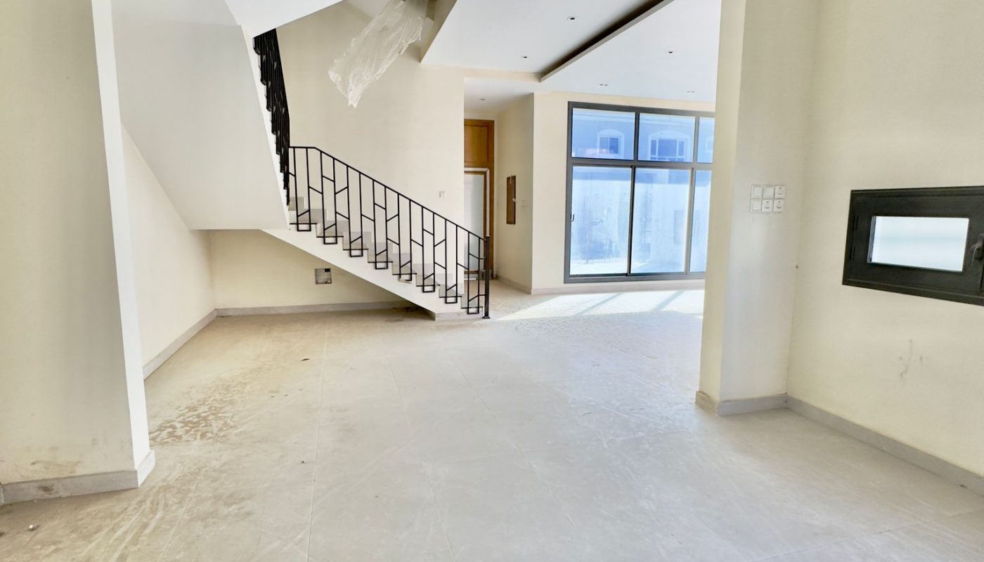 Unfurnished, modern interior space with white walls, tiled floor, a staircase with black railing, and large windows letting in natural light.