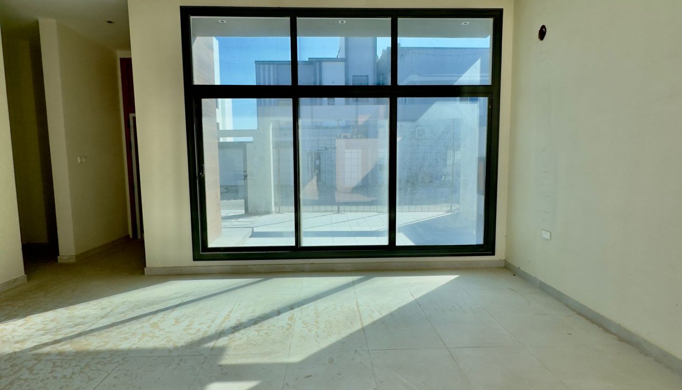 Empty room with large floor-to-ceiling window, beige tiled floor, cream walls, and sunlight casting shadows inside.