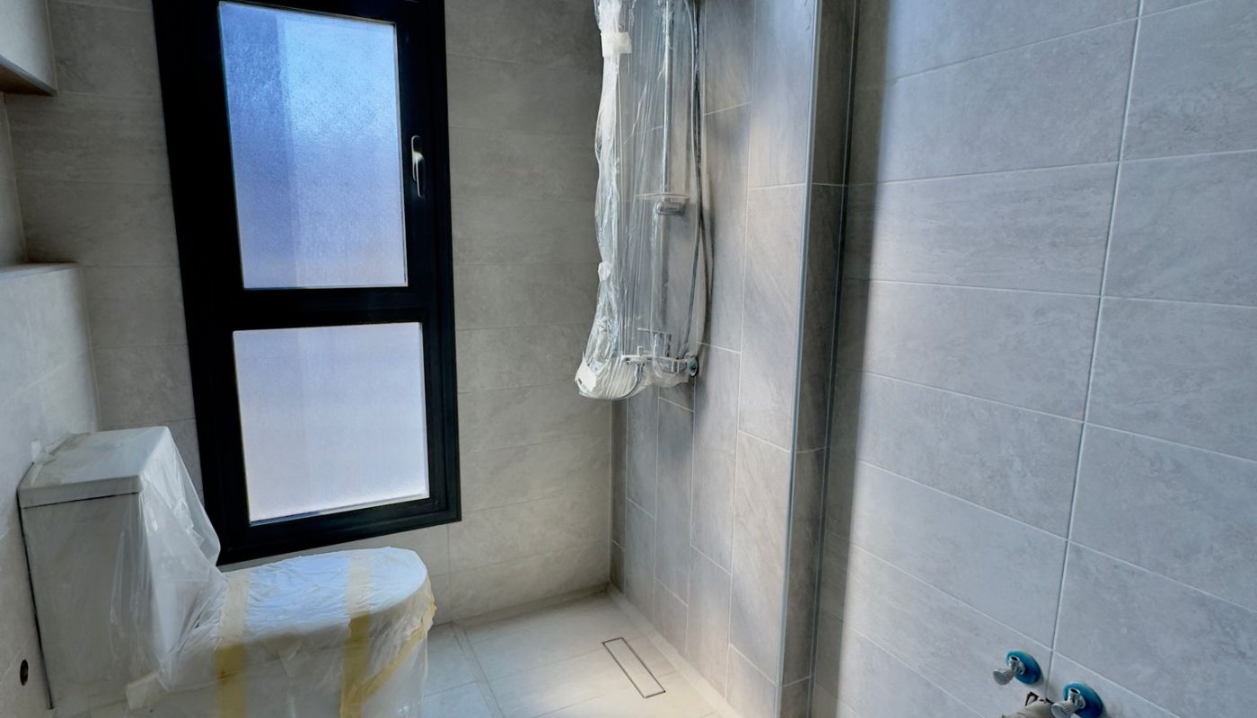 A bathroom with gray tiled walls and floor, a window, an uncovered toilet and shower fixture both wrapped in protective plastic, and exposed plumbing on the right wall.