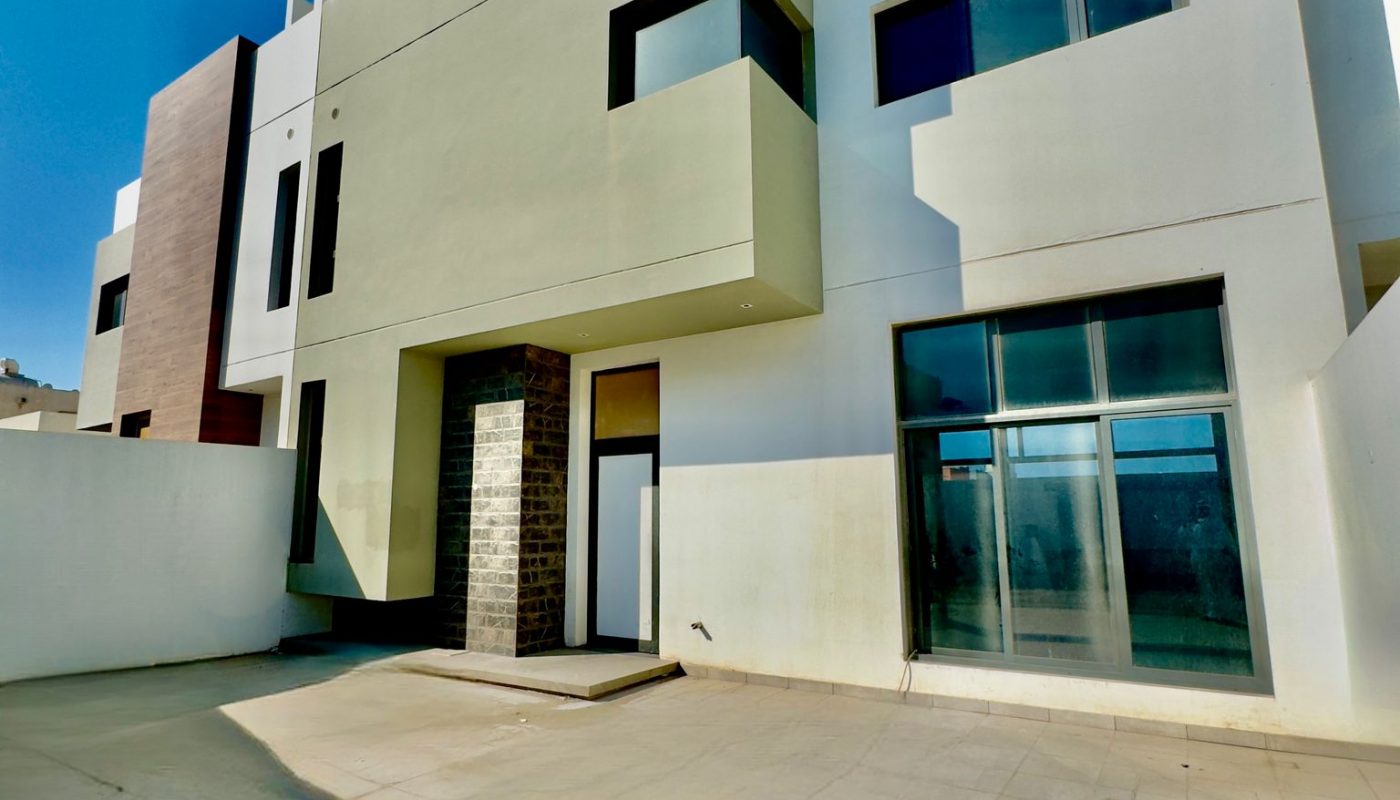 Modern two-story house with white and beige exterior, large windows, and a spacious tiled patio under a clear blue sky.