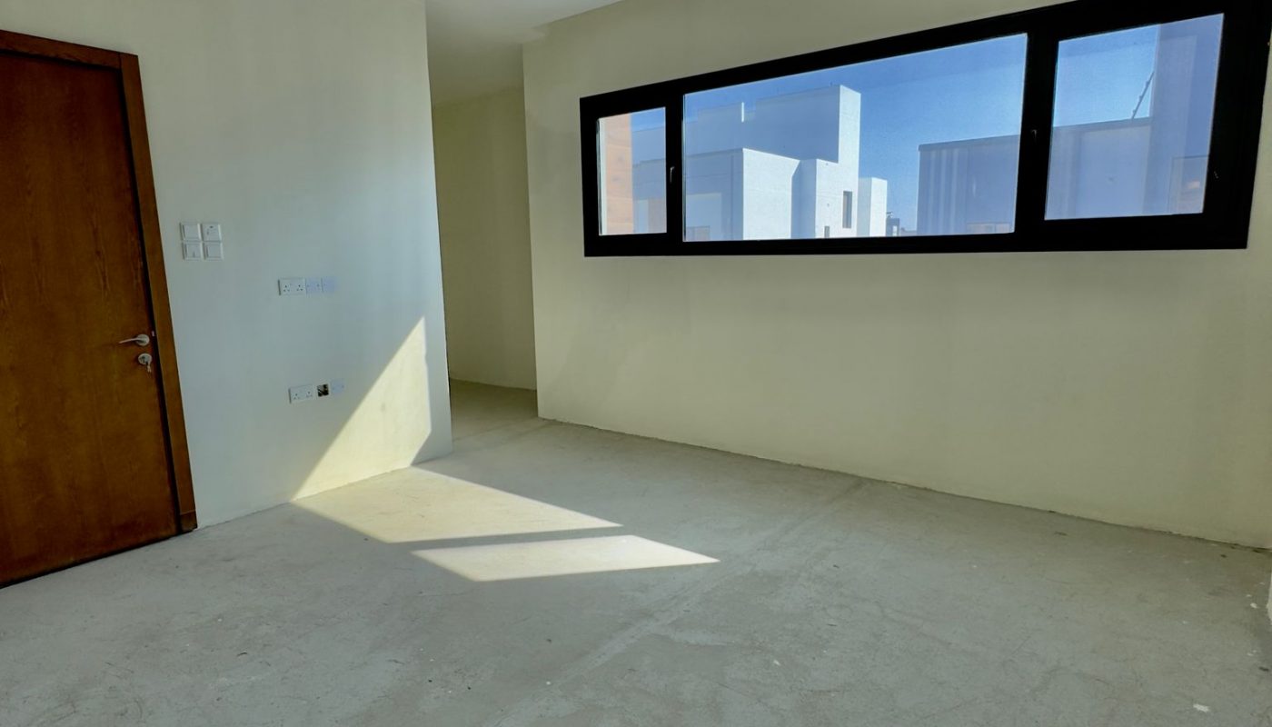 Empty room with a large window, white walls, concrete floor, wooden door on the left, recessed ceiling, and sunlight streaming in.
