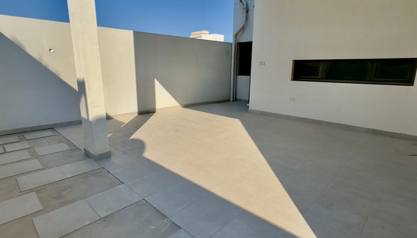 A modern outdoor patio with tiled flooring, white walls, a shaded area from the roof, and clear blue sky overhead.