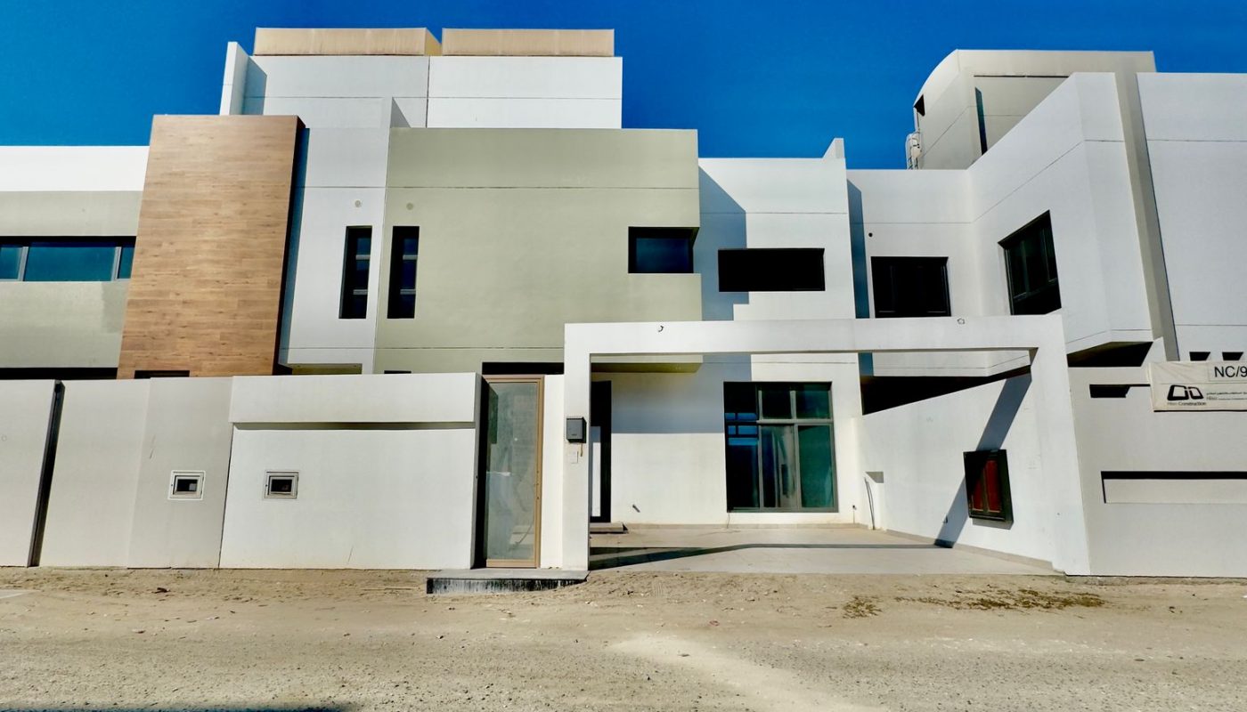 Modern white multi-story building with geometric design elements, large windows, and a sandy ground in front, under a clear blue sky.