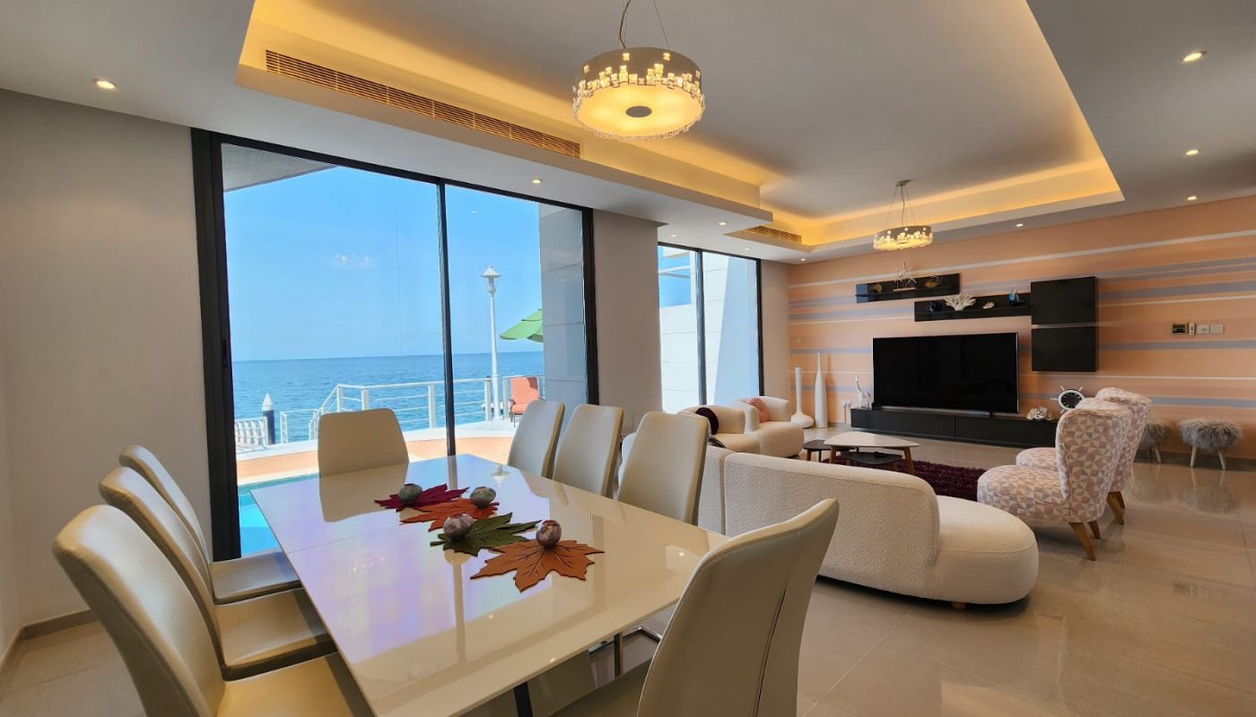Modern living and dining area with white furniture, large windows overlooking a pool and the sea, flat screen TV, and contemporary lighting.