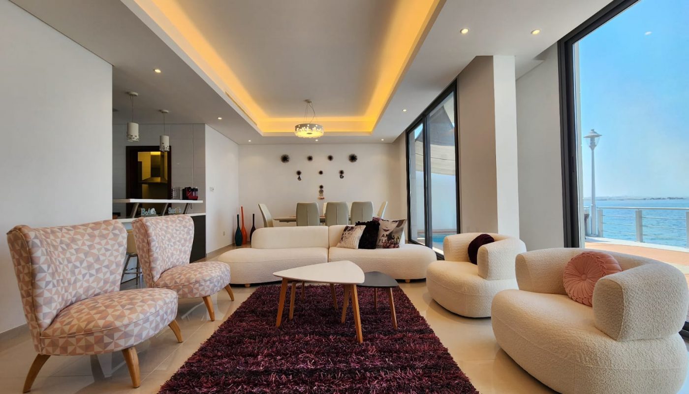 Modern living and dining area with contemporary furniture, large windows overlooking water, and a purple rug on a light tile floor.