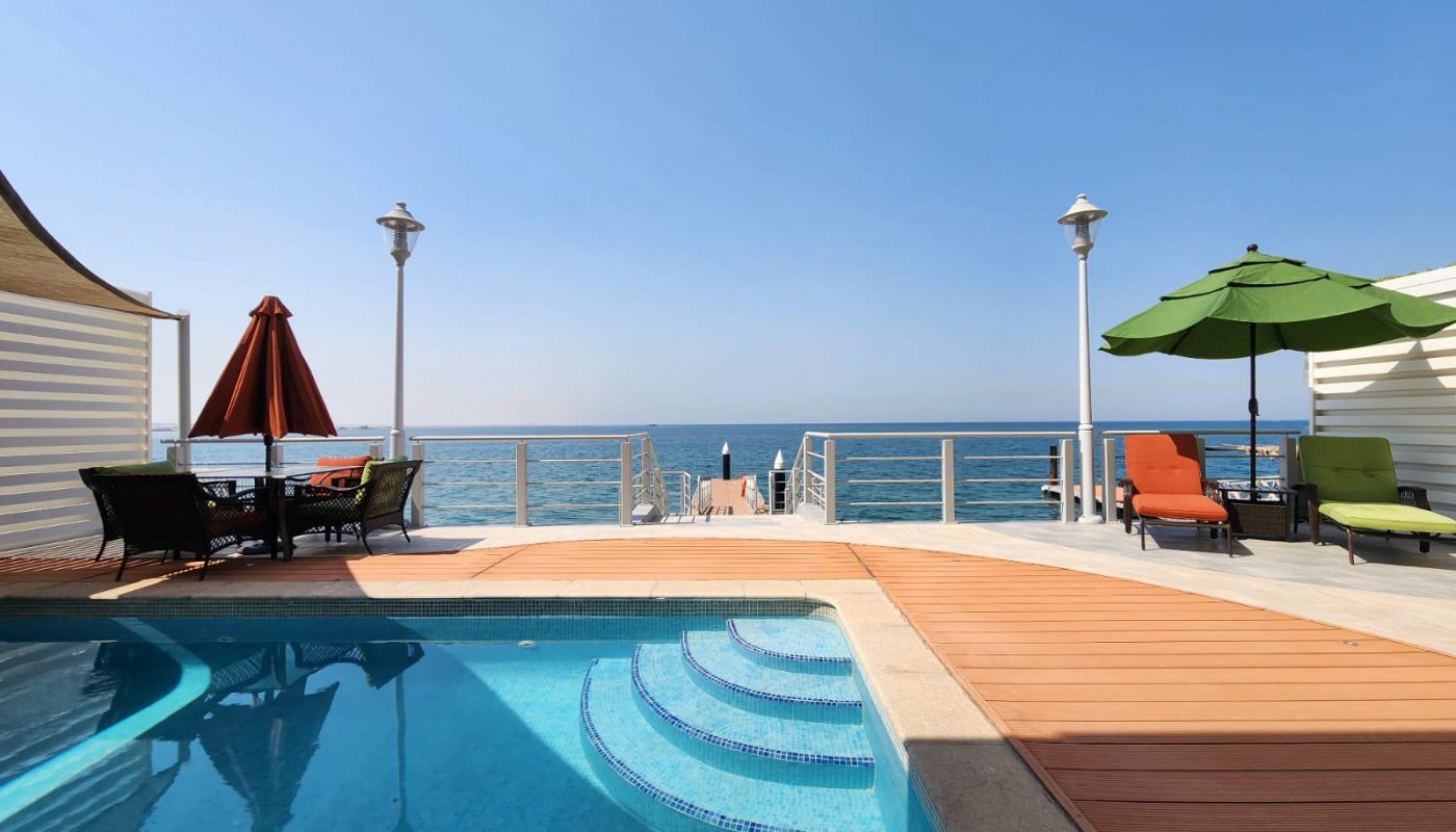 A poolside deck with lounge chairs, umbrellas, and a table overlooks the ocean under a clear blue sky.