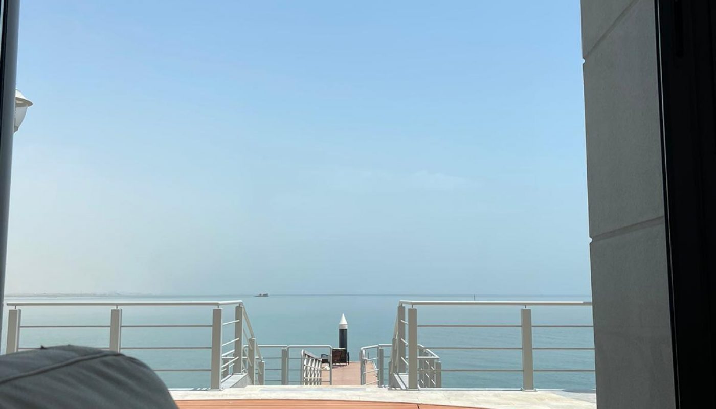 View from indoors of a white couch, looking out through a large window at a pool, wooden deck, and a dock extending over calm water under a clear sky.