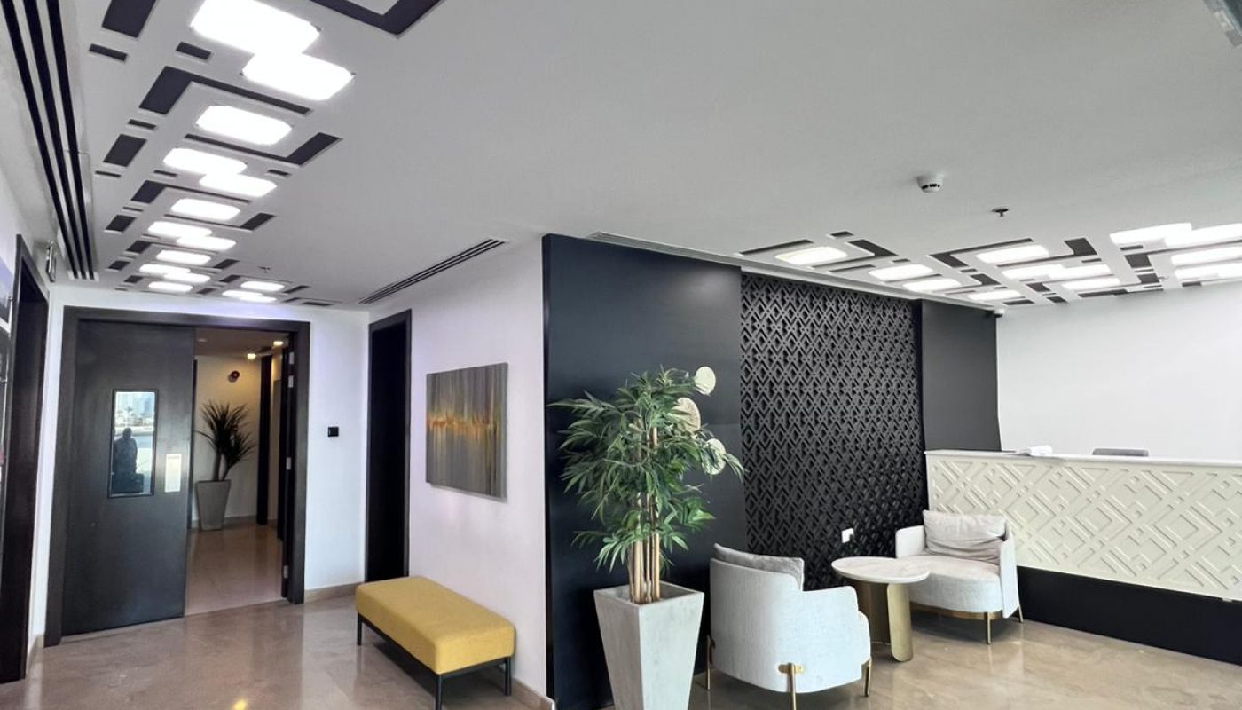 Modern office lobby with marble floor, a reception desk, potted plant, two chairs, a small table, yellow bench, and geometric ceiling lights.