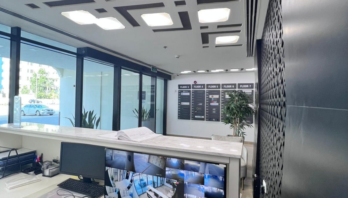 A modern office workspace with desks, chairs, plants, and a monitor displaying multiple security camera feeds. Large windows line one wall and information boards are visible in the background.
