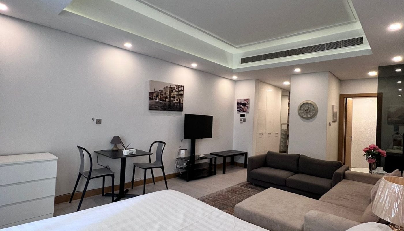 Modern hotel room with a large bed, sitting area with a sofa and TV, small dining table with chairs, white drawers, and recessed lighting. Walls are decorated with framed art.