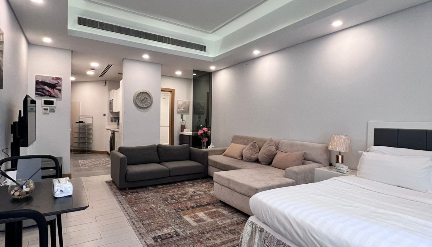 Modern studio apartment interior with a large bed, gray sofa, and patterned rug. Features a small dining area, open kitchen, and bright lighting. Walls and ceiling are white with recessed lighting.