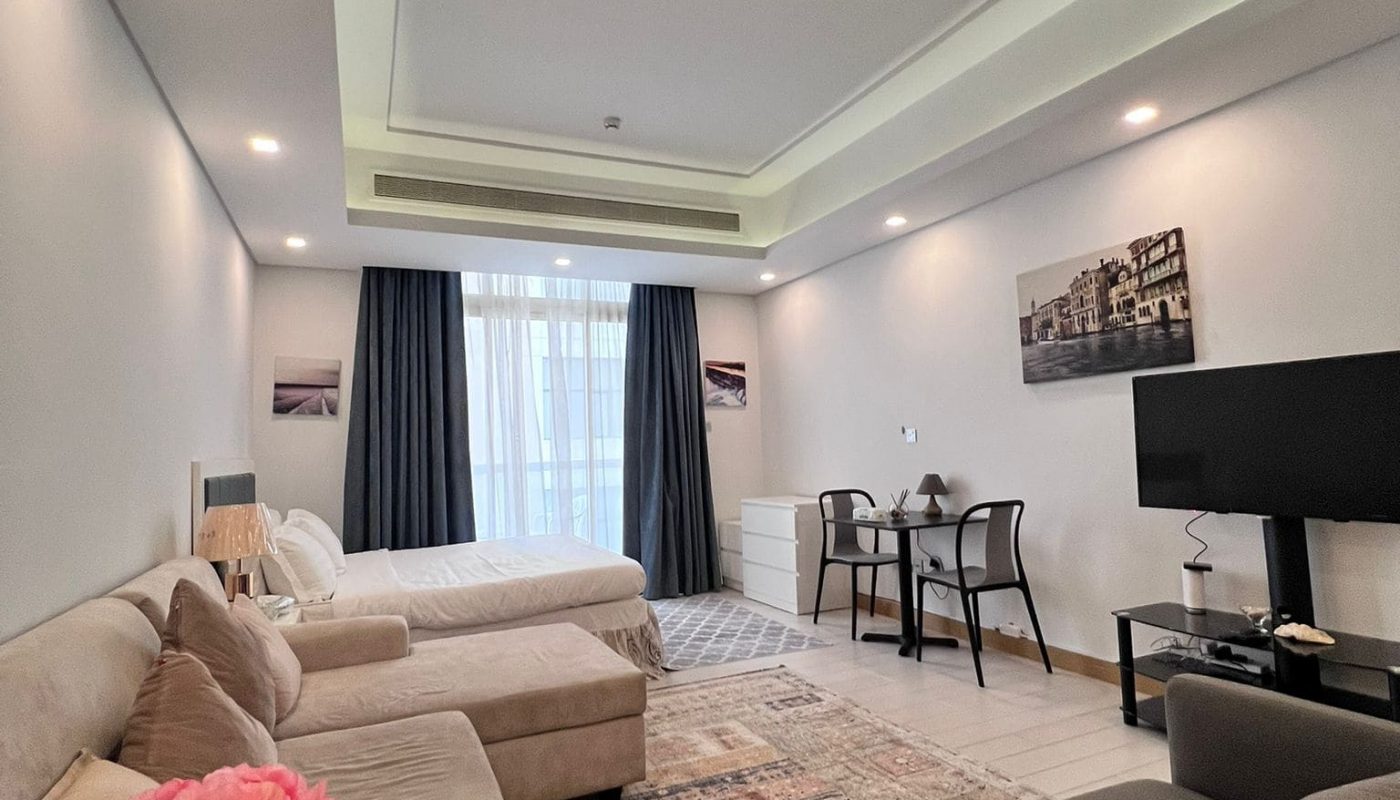 Living room with a bed, beige sectional sofa, pink flowers on table, TV, dining table with chairs, and large window with dark curtains. Walls have framed photos and ceiling has recessed lighting.