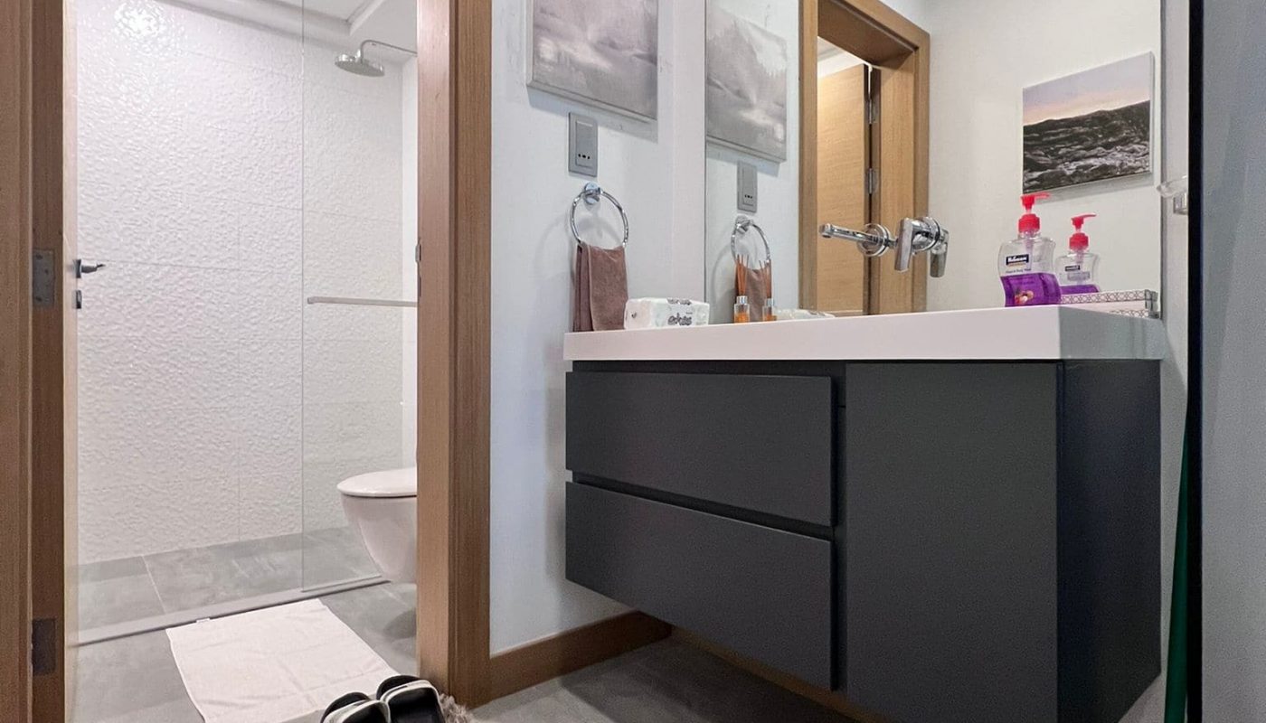 Modern bathroom with a floating vanity, large mirror, and walk-in shower. A brown rug and black slippers are on the floor. Wall art decorates the walls. Bright, overhead lighting.