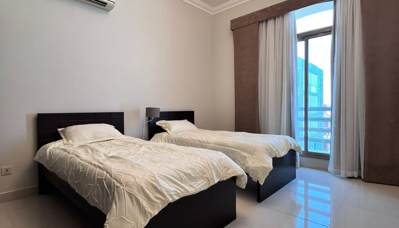 Two single beds with white bedding in a minimal bedroom. A window with curtains is on the right. A ceiling light fixture is above, and an air conditioning unit is on the wall.