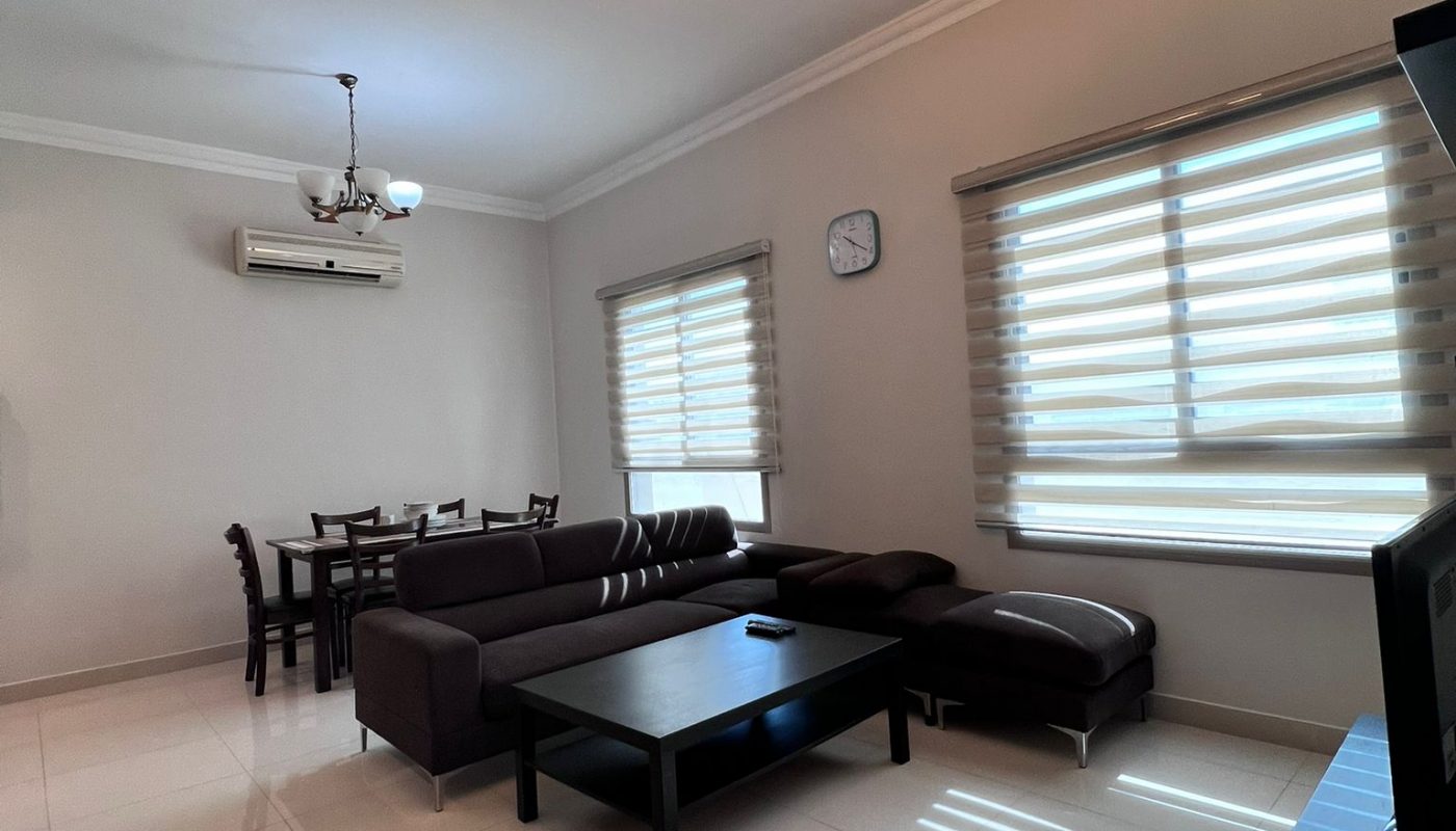 A modern living room with a dark sofa set, coffee table, and dining area in the background. Two windows have blinds, and there's a ceiling light fixture.