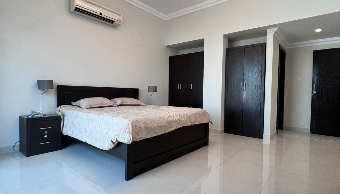 Minimalist bedroom with a double bed, bedside table, and lamps. White walls, tile floor, built-in wardrobes, ceiling light, and an air conditioner are visible.