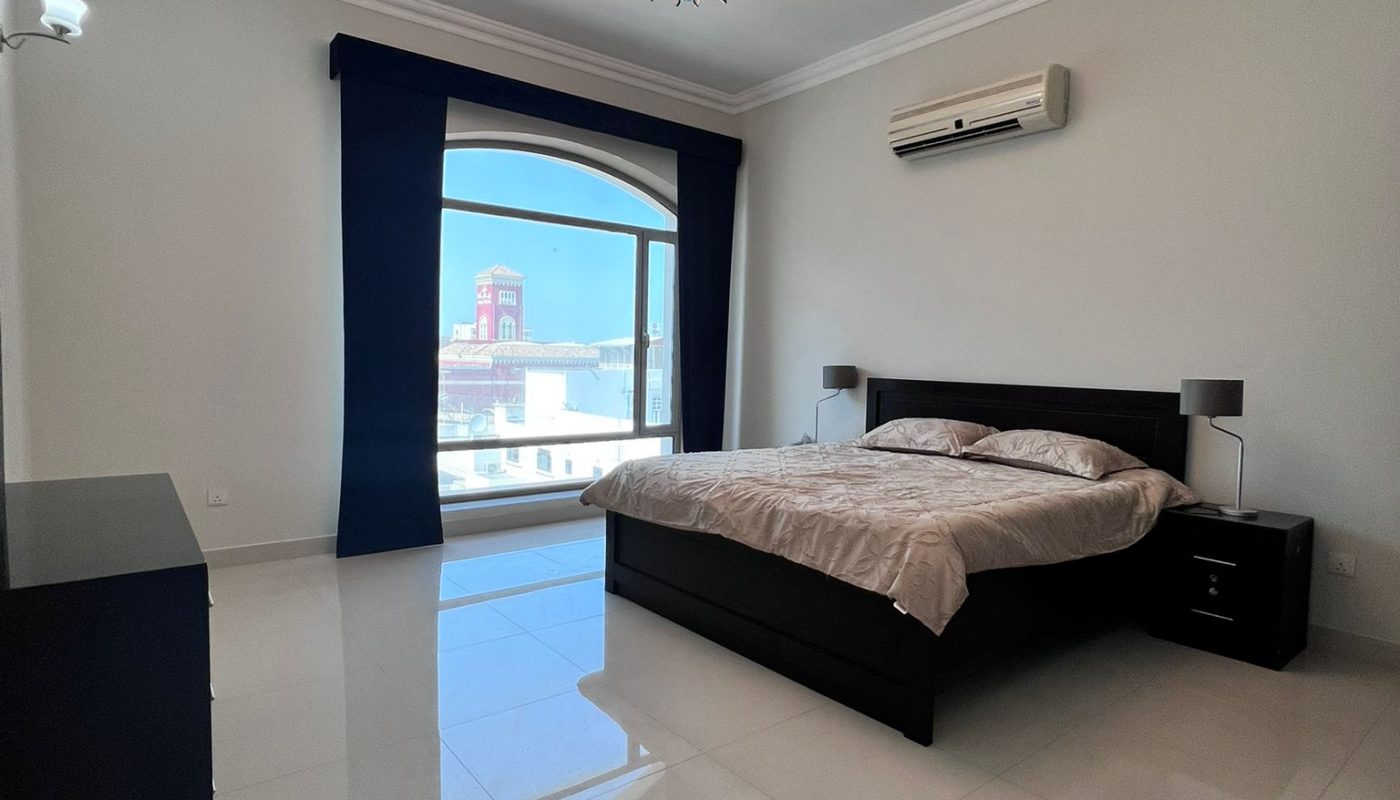 A modern bedroom with a large bed, two bedside tables, a window with blue curtains, a ceiling light fixture, and an air conditioning unit on the wall.
