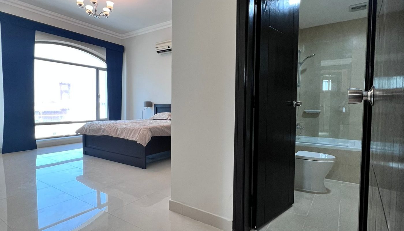 A bedroom with a large window, bed, and chandelier. An open door reveals a bathroom with a shower and toilet. The room has tiled floors and white walls.