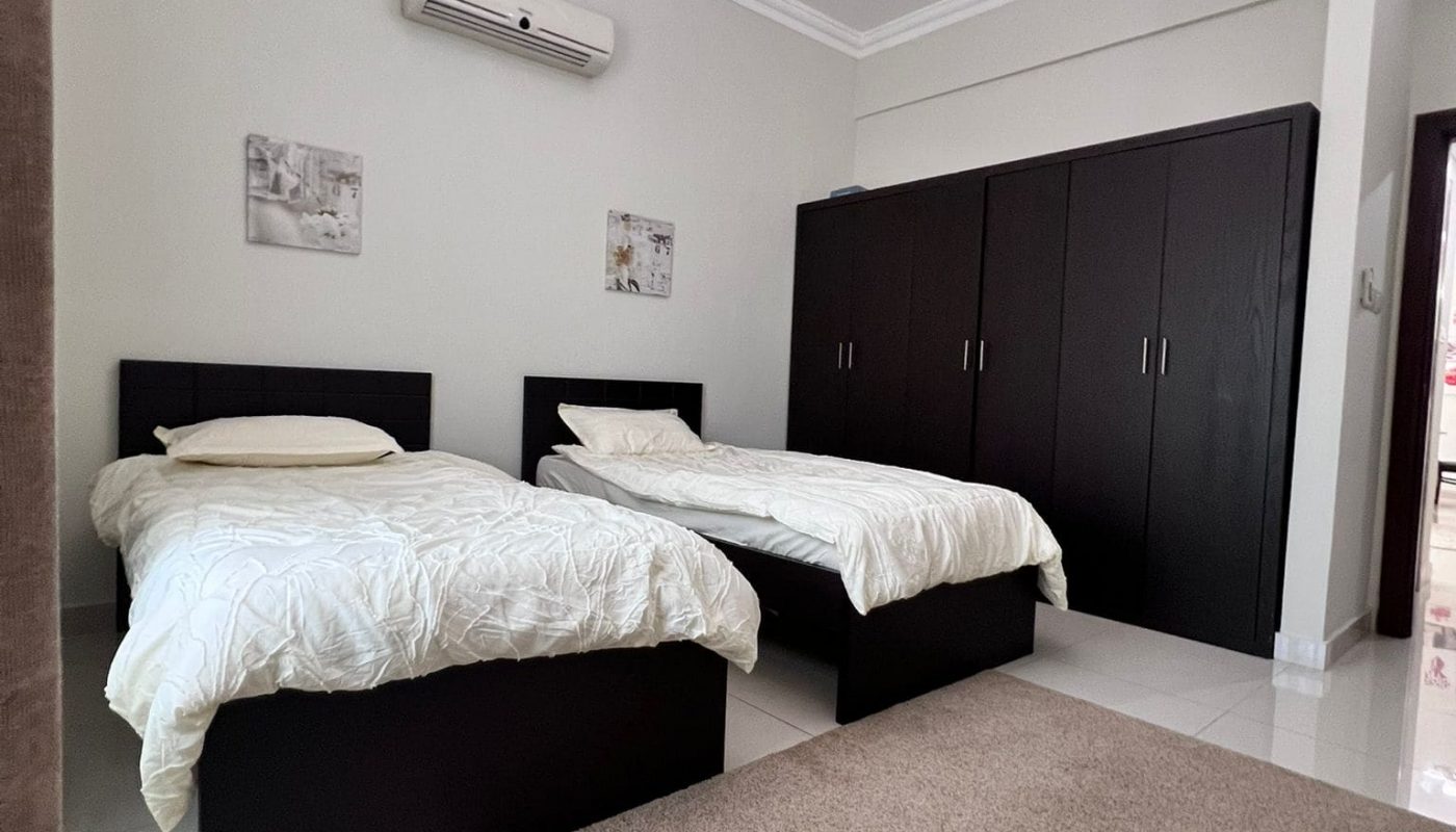 Bedroom with two single beds, white bedding, a brown rug, dark wardrobes, a ceiling light, and a wall-mounted air conditioner. Walls are light-colored with framed art.