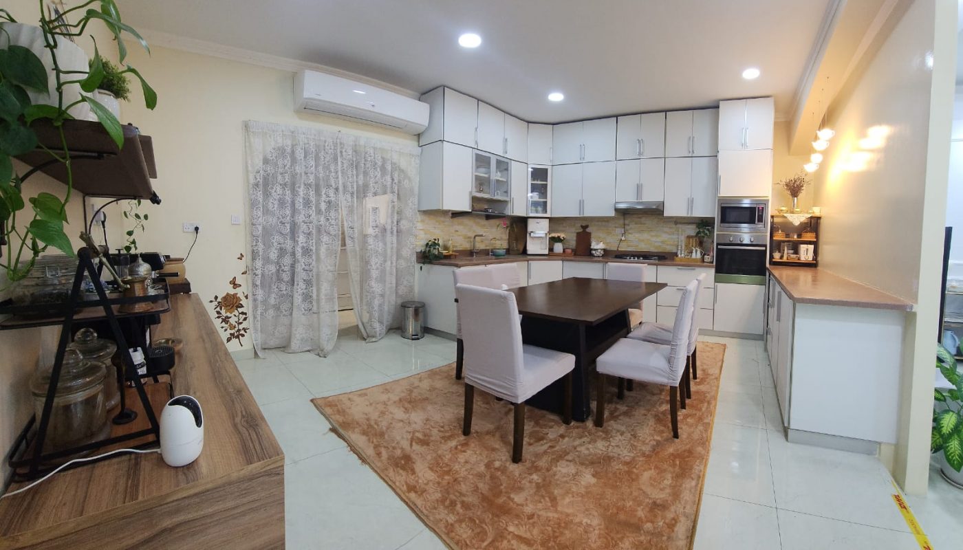 Modern kitchen with white cabinets, dining table with four chairs, brown rug, and plants. A curtain covers a doorway, and the room is well-lit with ceiling lights.
