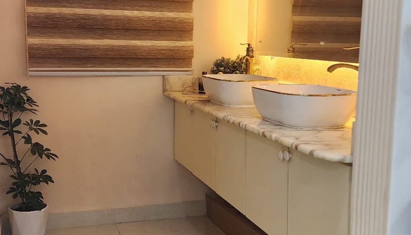 Modern bathroom with a large mirror, vessel sink on a marble countertop, potted plant, and a decorative rug on tiled floor. A window with a striped blind is on the left.