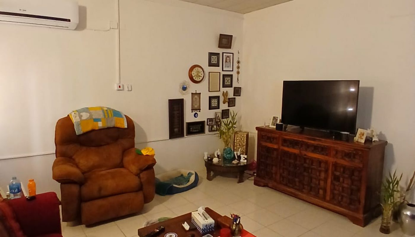 A living room with red sofas, a brown recliner, a wooden coffee table, TV on a cabinet, and framed photos on the wall; plants and decorative items are visible.