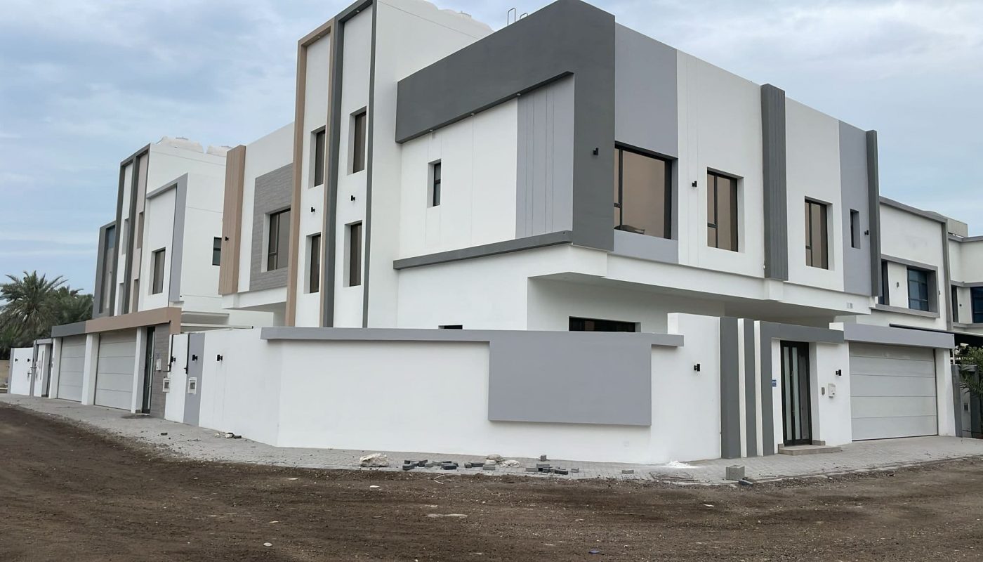 Modern two-story house with a flat roof, featuring a white and gray exterior. The area around the house has a dirt road and a small paved sidewalk.