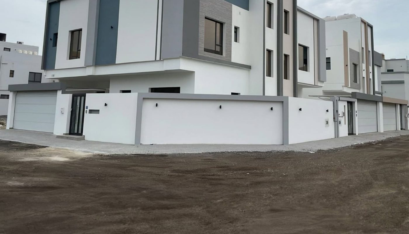 Modern two-story building with a minimalist design, featuring gray and white exterior walls, large windows, and a flat roof, surrounded by a paved area and a white fence.