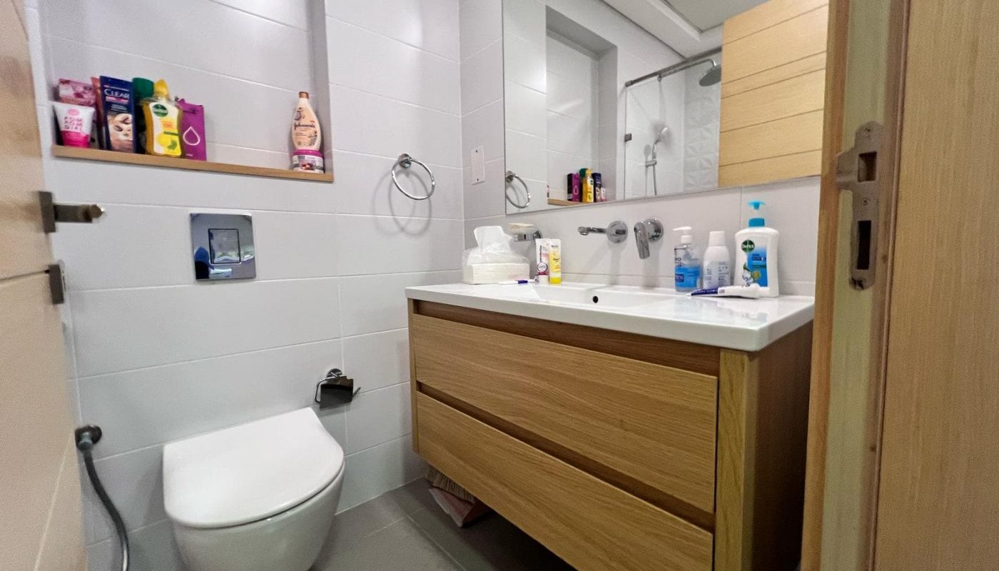 Modern bathroom with a wall-mounted toilet, wooden vanity with two drawers, and a sink. Various toiletries and cleaning supplies are on the countertop and shelves.