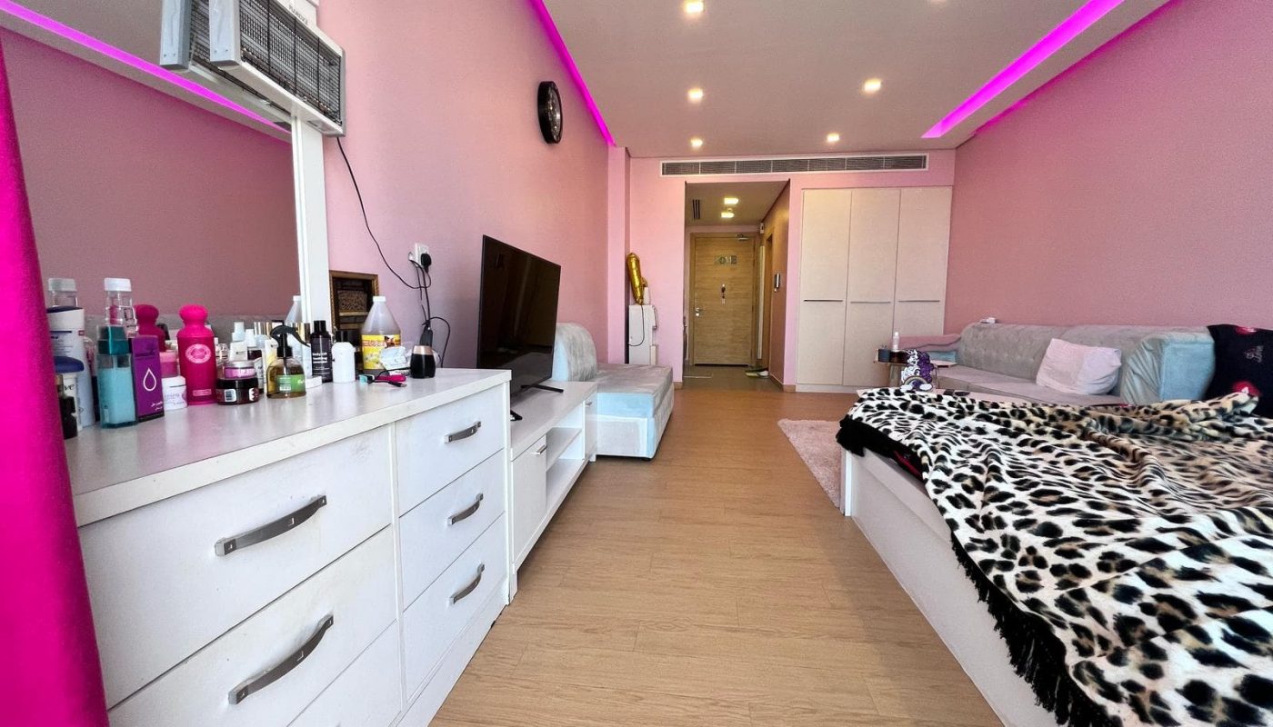 A bedroom with pink walls, featuring a bed with a leopard-print blanket, a dresser with various items, a TV, and a purple accent light strip on the ceiling. A door is seen in the background.
