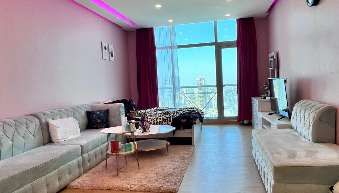 A modern living space with pink walls, a grey sectional sofa, a bed, a TV, and a balcony window with a city view. Purple curtains and ceiling lights add accent to the room.