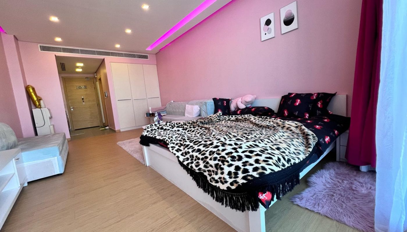 A pink-themed bedroom with a bed, leopard print blanket, artwork on the wall, couch, and wood flooring. Curtains and mood lighting enhance the ambiance.