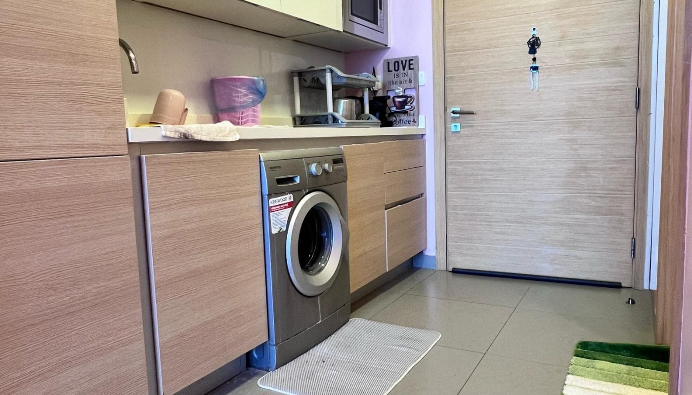 A compact kitchen features wooden cabinets, a washing machine, sink, and various kitchen items. A door with "HOME" decoration and a green rug are visible.