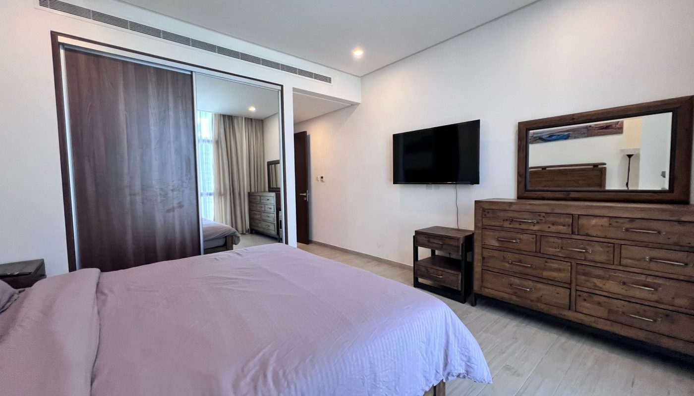 A bedroom with a neatly made bed, wooden dresser with mirror, wall-mounted TV, closet with sliding doors, and light wood flooring.