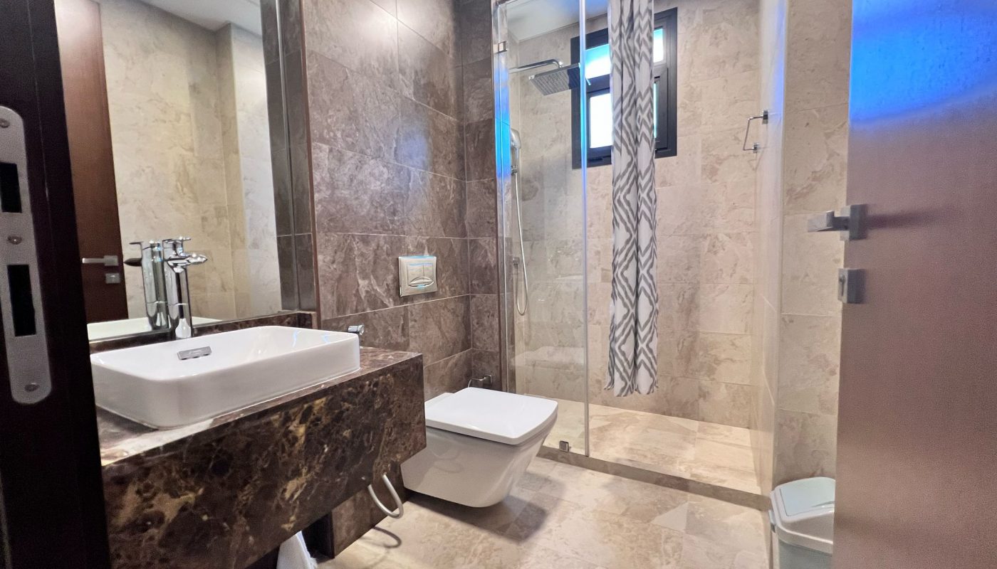 Modern bathroom with marble walls and floors, a sink, wall-mounted toilet, glass-enclosed shower, patterned shower curtain, and a small window.