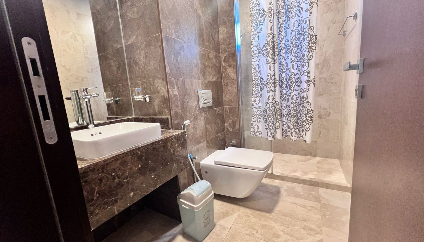 Modern bathroom with marble walls and floor, a floating sink, a wall-mounted toilet, a small trash bin, and a shower area with a patterned curtain.