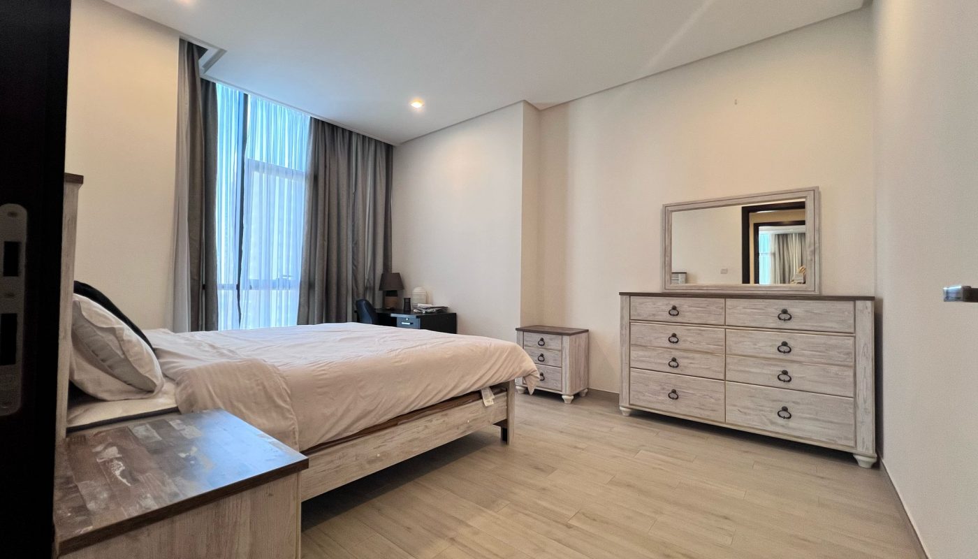 A bedroom with a bed, bedside table, dresser with mirror, chest of drawers, desk, and a large window with curtains. Light wood flooring and neutral walls.