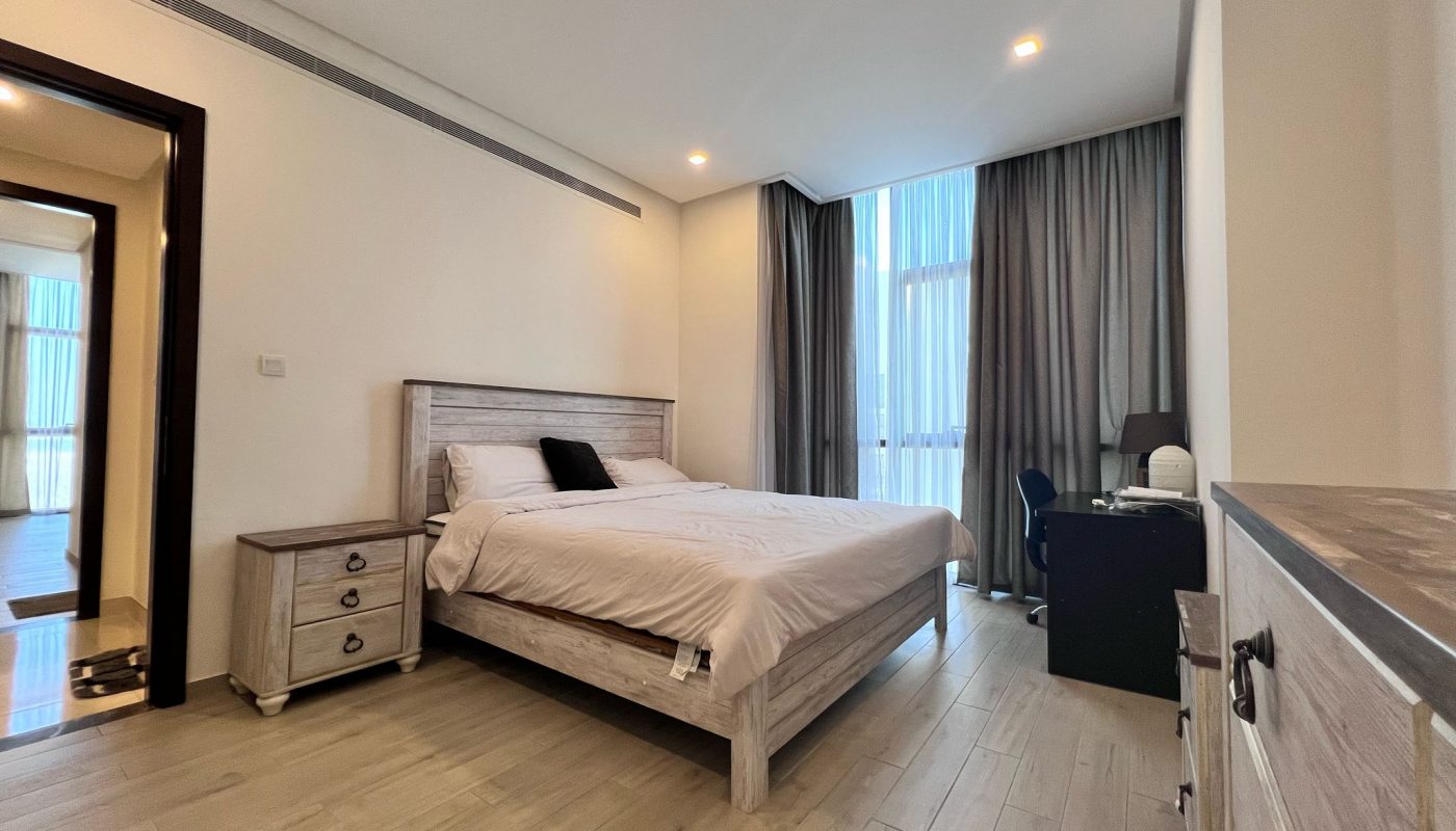 Modern bedroom with a double bed, bedside table, desk with a chair, and floor-to-ceiling window with curtains. Light wood flooring and neutral-colored walls.