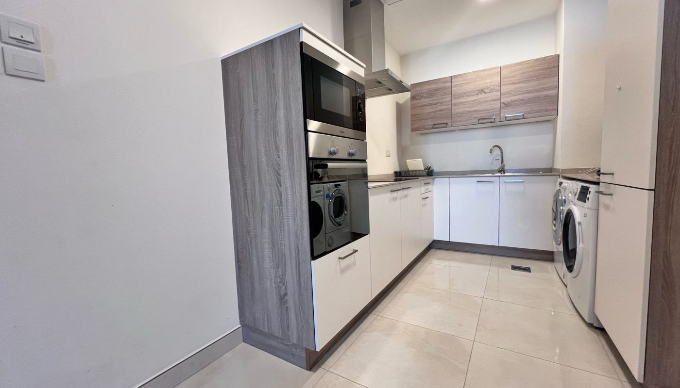 Modern kitchen with light-colored cabinets, built-in oven and microwave, stovetop with hood, sink, washing machine, and tile flooring.