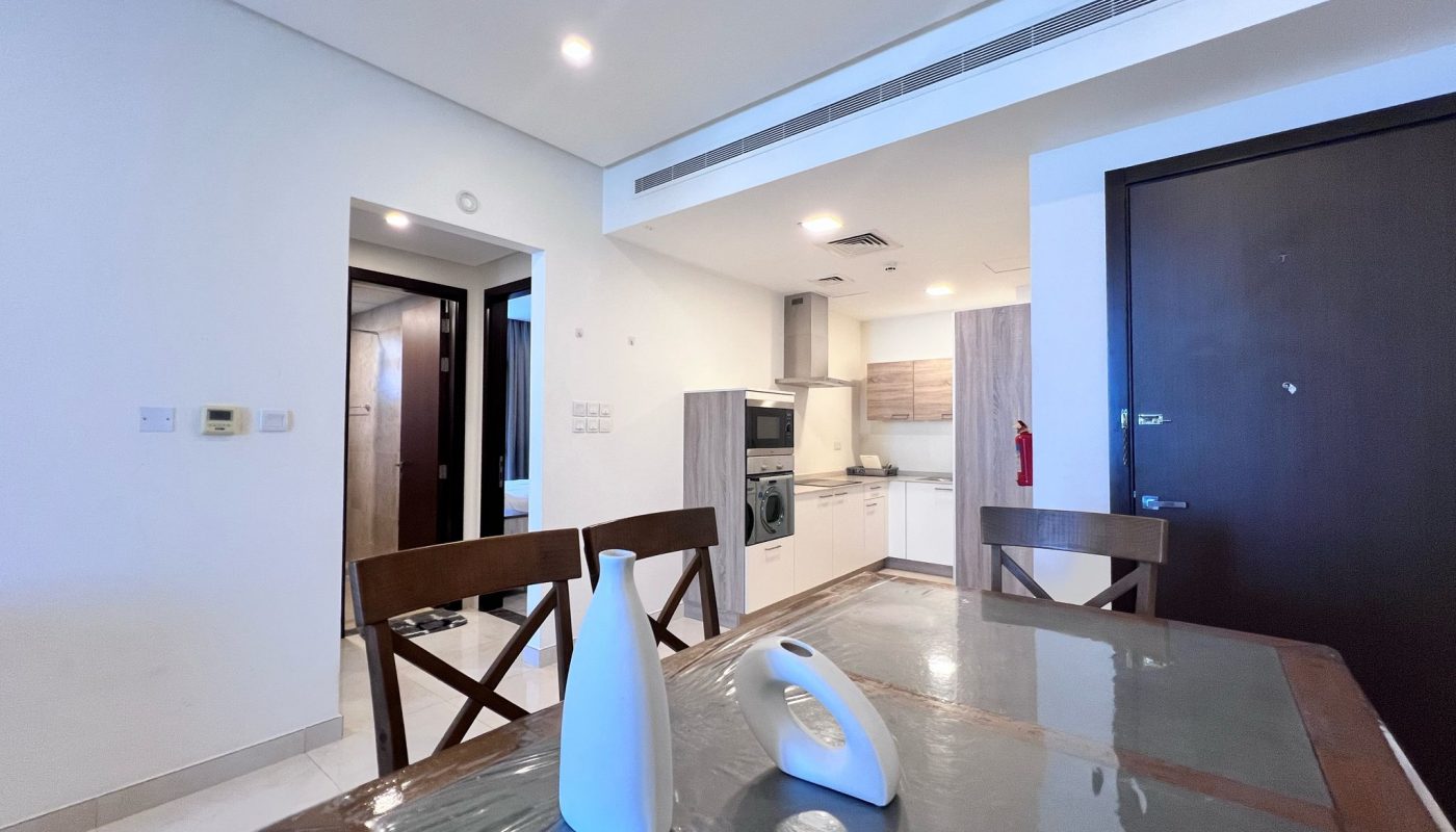A dining table with two white vases is in the foreground; a kitchen with light wood cabinets and built-in appliances is visible in the background.