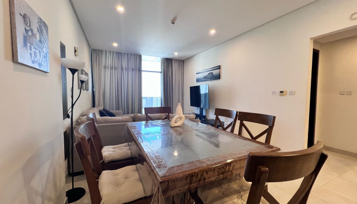 A dining area with a wooden table and six cushioned chairs, a living area with a TV, and large window with curtains in a modern apartment.