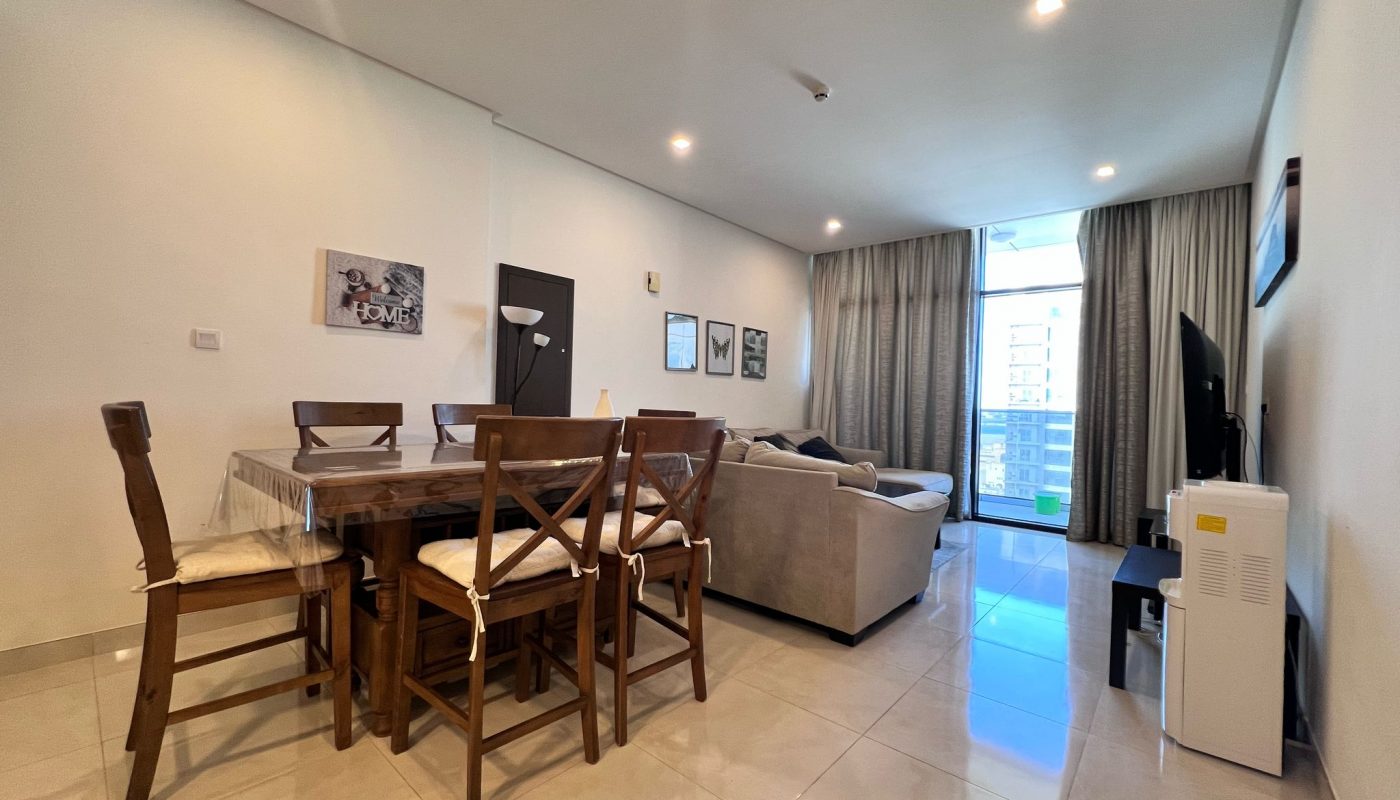 Modern living and dining area with a wooden table and chairs, beige sofa, wall-mounted TV, large window with curtains, and tiled floor.