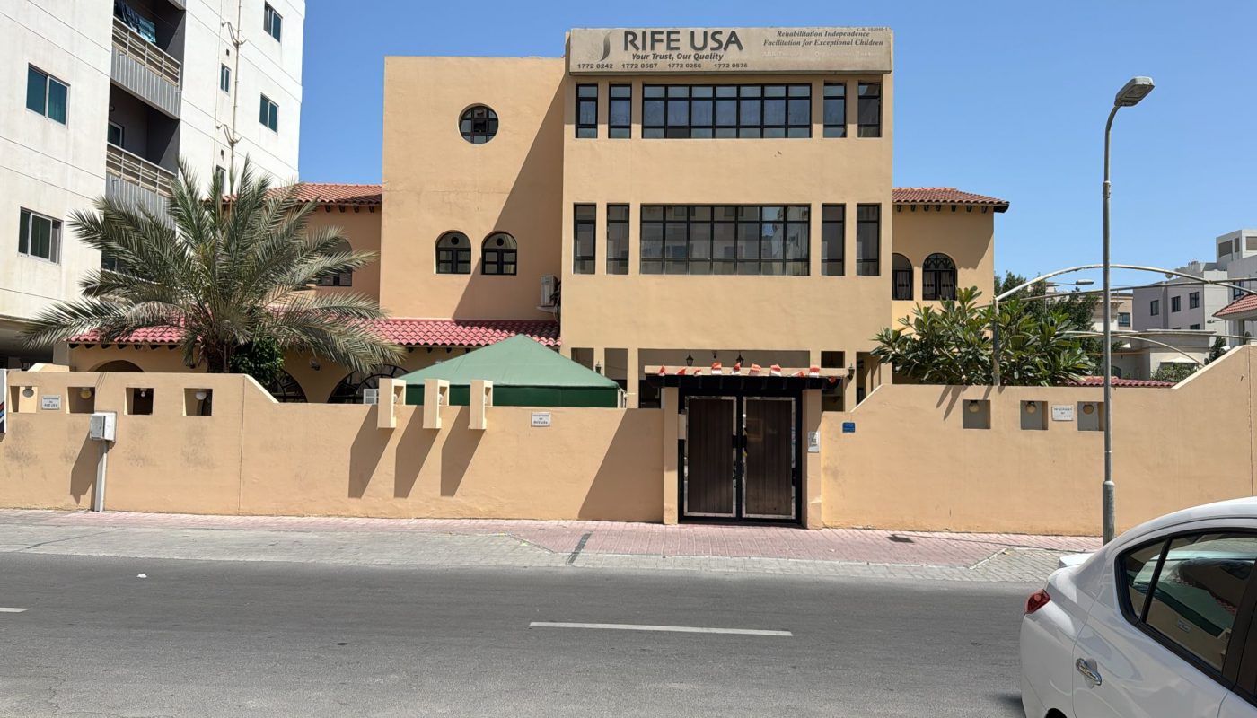 A tan building labeled "RIFE USA" with a fenced yard and palm tree is seen from across a quiet street on a sunny day.