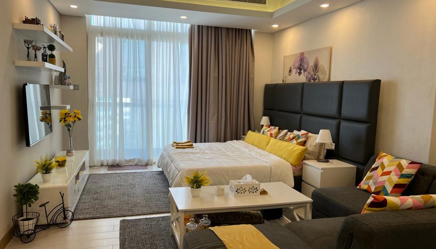 Modern bedroom with a double bed, sofa, TV, shelves with decor, and large window with sheer curtains. Bright accents include yellow pillows and flowers.
